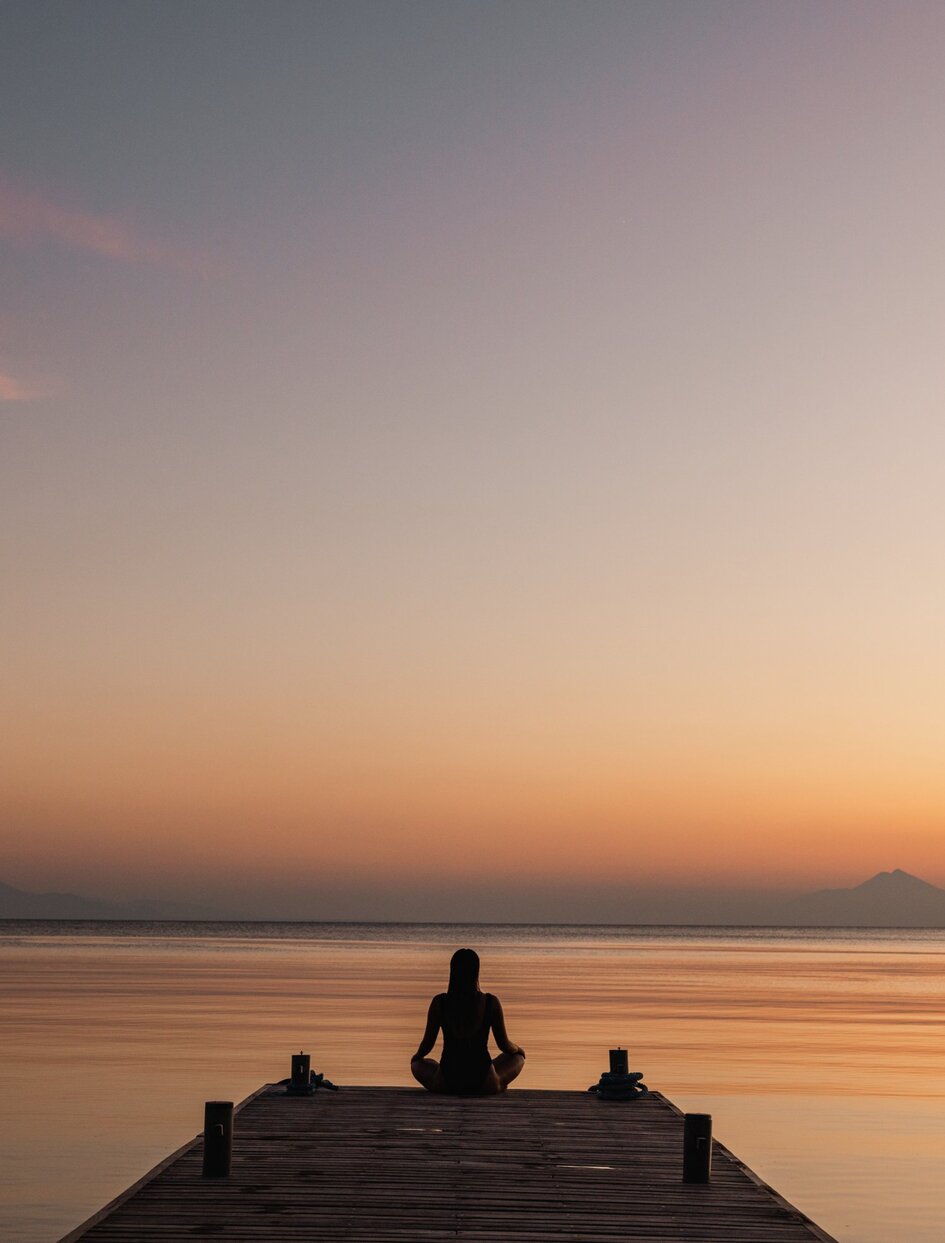 Solitary figure meditating on a wooden platform at Amanwana as the sun sets over calm waters.