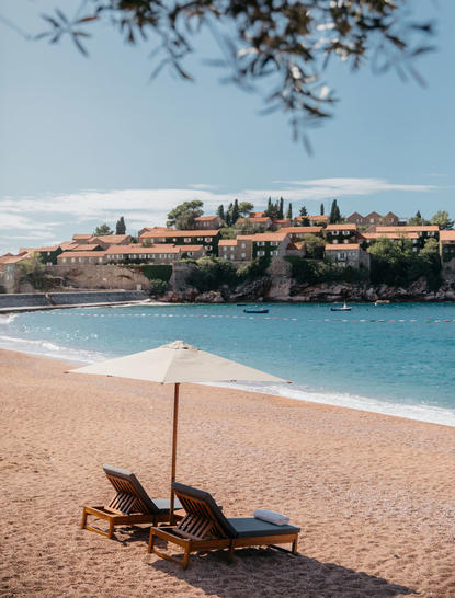 Wooden sunlounger with parasol on sandy beach at Aman Sveti Stefan, turquoise waters beyond.