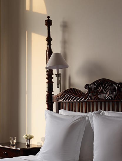 Wooden carved bedpost beside white linens at Amangalla, with candlelight casting shadows on the wall.