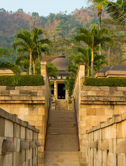 Pathway leading to a stone gateway at Amanjiwo resort, flanked by palm trees and terraced gardens.