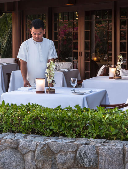 Staff member arranging table settings at Amanpulo's dining clubhouse, with stone terrace and tropical greenery in foreground.