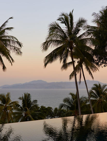 アマンプリ リゾートの9ベッドロームオーシャンヴィラから、夕暮れ時の海と山々を望む景観。ヤシの木が立ち、プール越しに静かな眺めが広がる。