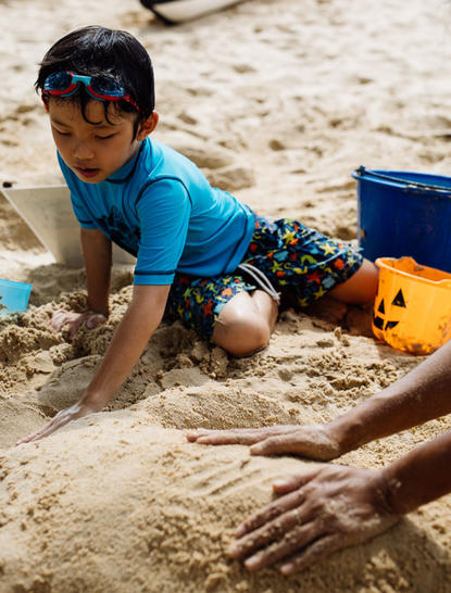 Bambino in maglietta blu gioca sulla spiaggia sabbiosa di Amanpuri, circondato da secchielli e giocattoli.