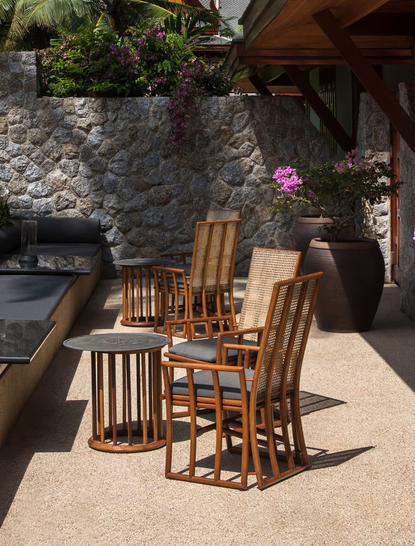 Wooden dining table and chairs set on a sunny terrace at Amanpuri, with stone walls and flowering plants nearby.
