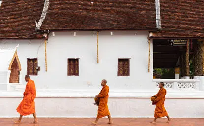 Buddhist monks in saffron robes walking past a whitewashed temple building at Amantaka, Laos.