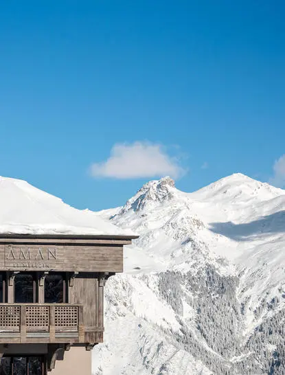Wooden chalet at Aman Le Mélézin with snow-capped Alpine peaks against clear blue sky.
