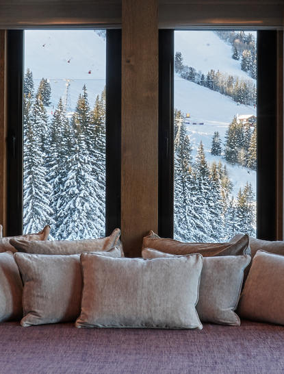 Bedroom windows at Aman Le Mélézin frame snow-covered Alpine forest views, with cushioned seating below.