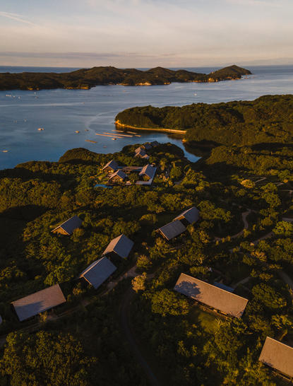 Wooden boardwalk through moss-covered rocks at Amanemu, with coastal waters and distant hills at dusk.