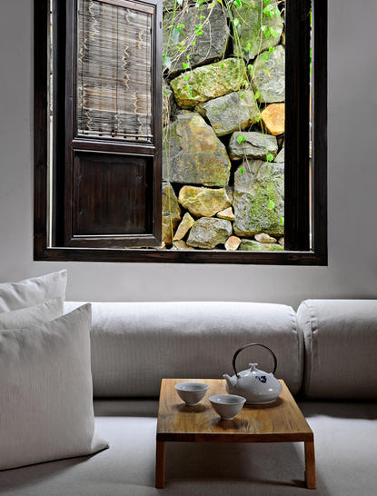 A living area at Amanfayun with a grey sofa, wooden table, and view through window to stone courtyard beyond.