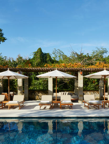 Poolside lounge area at Aman Villas at Nusa Dua with pergolas, seating, and mountain views.