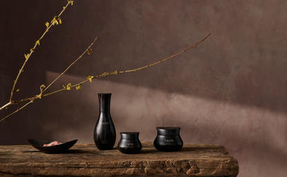 Aman Skincare campaign shot featuring dark glass bottles and vessels arranged on a wooden surface with dried branches in an office setting.