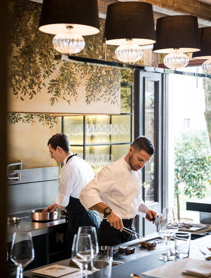Two chefs prepare a meal in the dining kitchen at Aman Venice, with pendant lights overhead and greenery framing the window.
