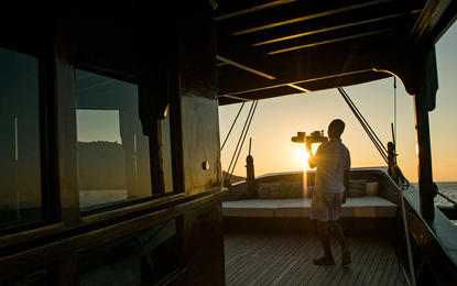 Warm sunset light floods a covered wooden deck at Amankila, where a figure stands gazing towards the horizon over water.