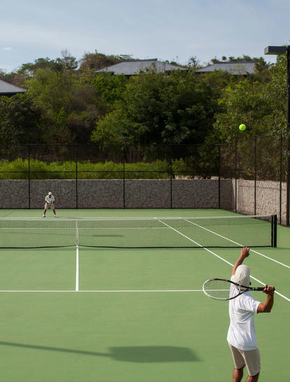Family member playing tennis on an outdoor court at Amanoi, with lush hillside vegetation beyond.