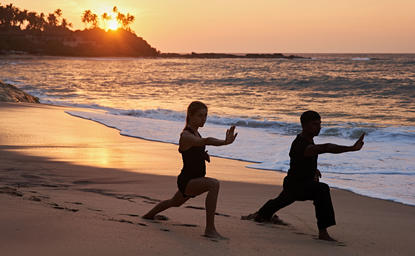 Two people practising yoga on Amanwella's beach at sunrise, with golden light reflecting on the sand and sea.