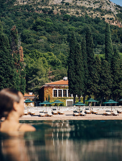 Waterfront view of a terracotta building nestled among cypress trees at Aman Sveti Stefan, with calm waters in the foreground.