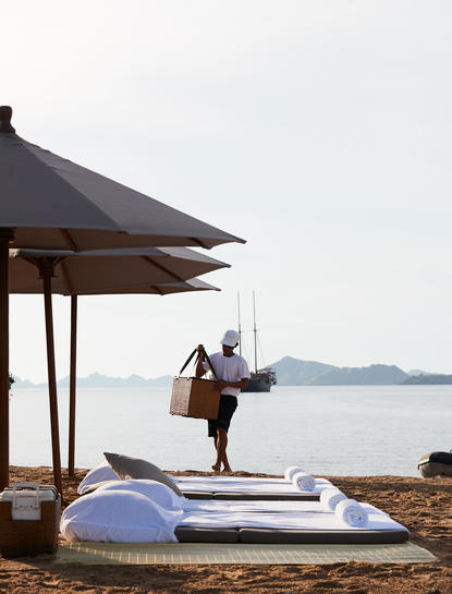 A figure in a wetsuit walks along the beach at Amandira, passing a sun lounger beneath a dark pavilion with views across calm waters.
