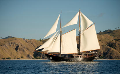 Traditional sailing vessel with cream sails on blue water, rocky coastline at Amandira beyond.