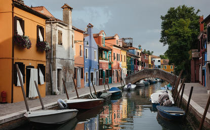 Colourful waterfront buildings reflected in a calm canal at Aman Venice, with a traditional wooden bridge connecting the vibrant structures along the water's edge.