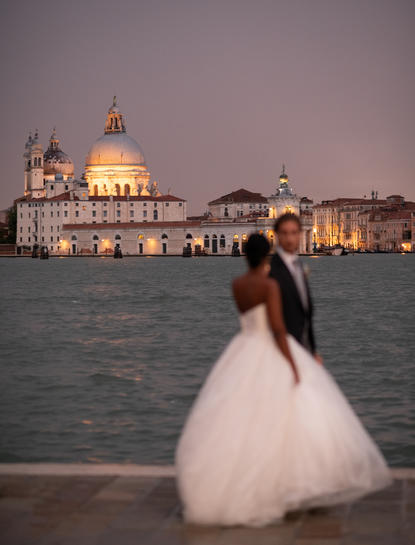 Bride in white gown gazing towards the illuminated Basilica di Santa Maria della Salute at dusk in Venice.