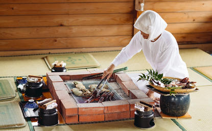 安缦伊沐度假村中，一位客人跪坐在木地板上，围绕着传统的烹饪工具和新鲜食材进行料理准备。