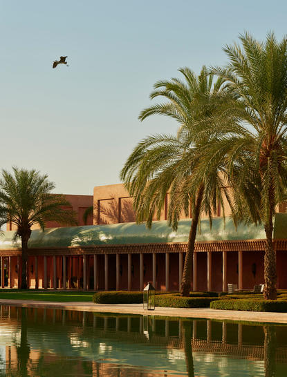 Palm trees line a tranquil waterway at Amanjena, with terracotta buildings reflected in still water.