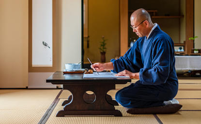 Man in blue robe seated on tatami mat at low desk, practising ink painting at Aman Kyoto resort.