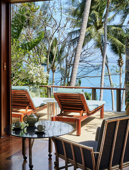 Drei-Zimmer-Strandvilla in Amanpuri mit Meerblick vom Wohnbereich, hölzerne Liegestühle und Palmen.