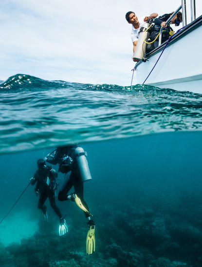 ডাইভার নীল সমুদ্রে স্নরকেল করছে, একটি নৌকা থেকে অন্য ব্যক্তি নজরে রয়েছে, আমানপুলো রিসর্টে।