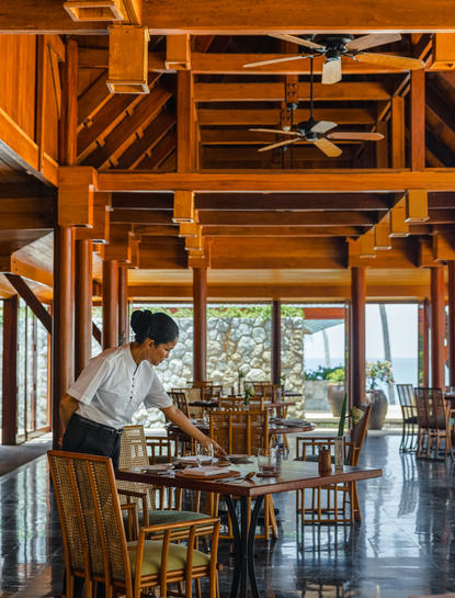 A guest seated at a wooden table inside a timber-framed dining pavilion at Amanpuri resort, Thailand.