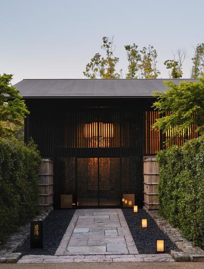 Dark timber entrance pavilion with illuminated windows at Aman Kyoto, framed by lush greenery.