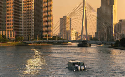 Aman Tokyo's private cruise vessel gliding through the Sumida River at golden hour, with illuminated skyscrapers lining the waterway.