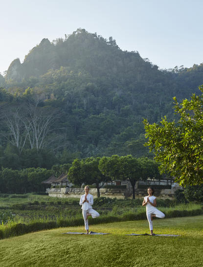 Deux femmes pratiquent le yoga sur une pelouse verdoyante face aux montagnes boisées d'Amanjiwo.