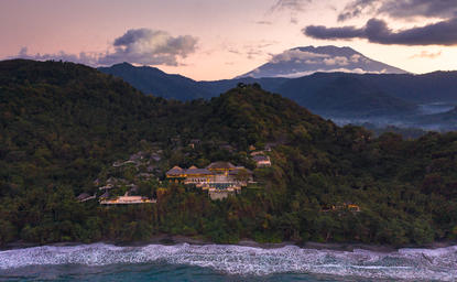 Aerial view of Amankila at dusk, with forested hills descending to a crescent beach and calm waters beyond.
