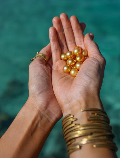 Hands holding golden pearls above turquoise water at Amanpulo resort.