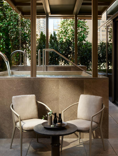 Spa House Terrace at Aman New York with two cream armchairs facing a small table, surrounded by verdant greenery and open glass walls.