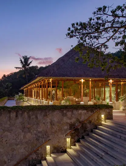 Amankila resort restaurant exterior at dusk, illuminated wooden structure with mountain backdrop.