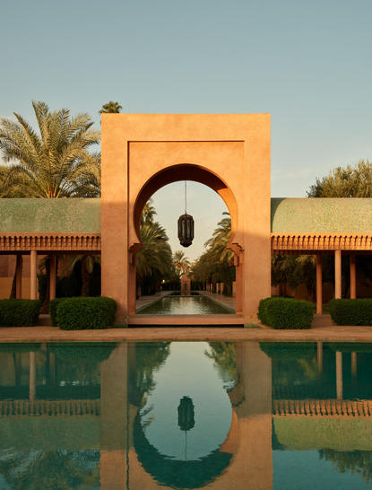 Amanjena's terracotta archway reflected in a still pool, with palm trees and pavilions either side.