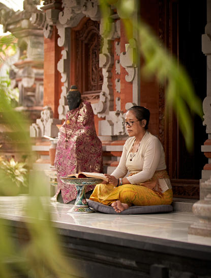 A woman sits cross-legged on a stone platform at Amandari resort in Ubud, Bali, surrounded by traditional Balinese numerology offerings and ornate temple architecture.