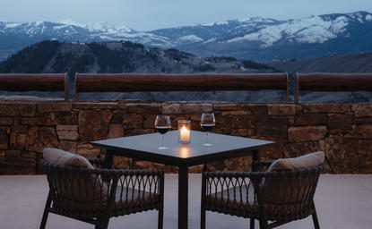 Outdoor dining setup at Amangani with two wicker chairs flanking a round table, snow-capped mountains visible in the distance at dusk.