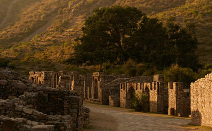 Gateway and tree at Amanbagh, with village ruins and hills beyond in golden evening light.