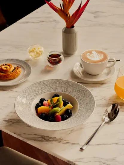 Amanera breakfast table with fresh fruit bowl, pastry plate, and cappuccino on marble surface.