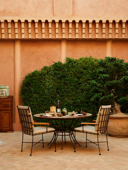 Dining table and chairs on terracotta courtyard at Amanjena, with green shrub and traditional ochre wall.