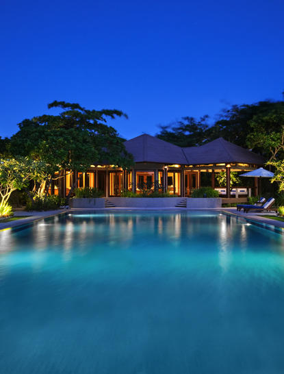 Amanpulo resort pool illuminated at dusk, turquoise water reflecting warm golden lights from pavilions beneath twilight sky.