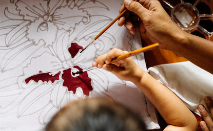 Hands creating an intricate batik pattern with deep red dye at Amanpuri resort.