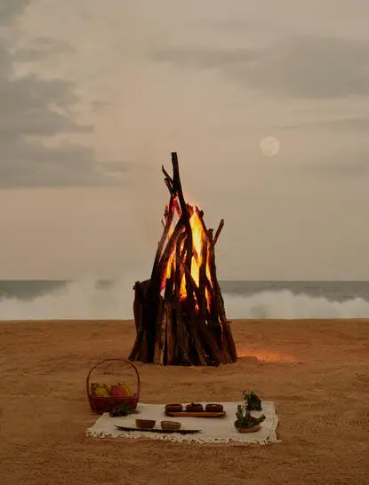 Feu de plage au coucher du soleil à Amanera, avec bois empilés sur le sable face à l'océan.