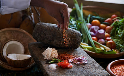 Hands preparing ingredients during a cooking lesson at Amanwella, Sri Lanka, with fresh vegetables and spices arranged on a stone surface.