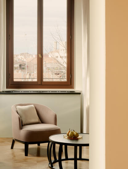 安缦威尼斯酒店浴室内，米色墙壁旁放置一把棕色扶手椅和小茶几，窗外可见威尼斯城景。