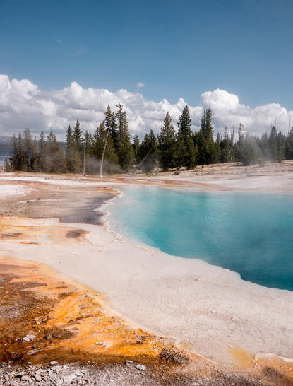 Turquoise hot spring with mineral deposits at Amangani's Yellowstone experience, evergreen forest and snow-capped mountains beyond.