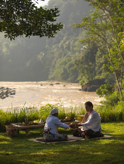 Two guests seated on grass overlooking a river at Amanjiwo, Central Java, during a wellness retreat.
